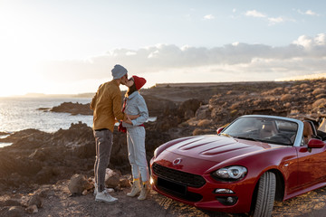 Young lovely couple kissing on the beach, traveling by car on the rocky ocean coast on a sunset....