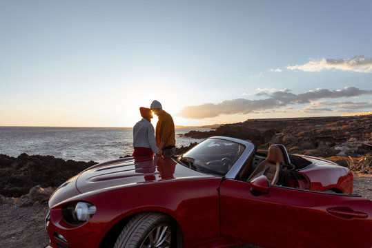 Couple enjoying beautiful views on the ocean, hugging together near the car on the rocky coast, wide view from the side with copy space on the sky