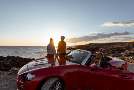 Couple enjoying beautiful views on the ocean, hugging together near the car on the rocky coast, wide view from the side with copy space on the sky