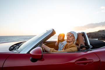Joyful couple enjoying vacations, driving together convertible car near the ocean on a sunset....
