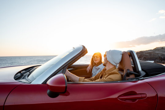 Joyful couple enjoying vacations, driving together convertible car near the ocean on a sunset. Happy vacation, love and travel concept
