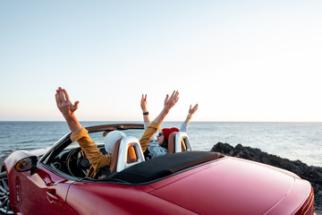 Couple driving convertible car, traveling near the ocean on a sunset, view from the backside. Happy...