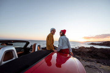 Portrait of a lovely couple dressed casually in hats sitting together on a car during a sunset, traveling near the ocean. Happy vacations and traveling by car concept