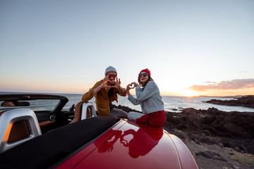 Portrait of a lovely couple dressed casually in hats sitting together on a car during a sunset,...