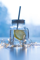 A jar with a straw filled with water and a lemon slice on a table with a light garland