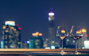 Two glasses of red wine on table of rooftop bar with colorful bokeh of city lights.