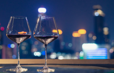 Two glasses of red wine on table of rooftop bar with colorful bokeh of city lights.