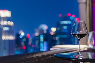 A glass of red wine on table of rooftop bar with colorful bokeh of city lights.