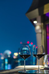 Two glasses of red wine on table of rooftop bar with colorful bokeh of city lights.