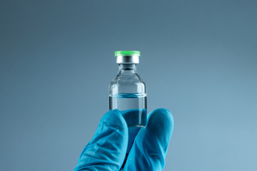 Hand in a blue glove holds a bottle with a vaccine close-up on a gray background, medical concept vaccination subcutaneous injection, dose. Disease treatment immunization.