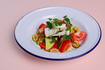 Traditional Greek salad with feta cheese and vegetables served in white plate over bright pastel pink background.