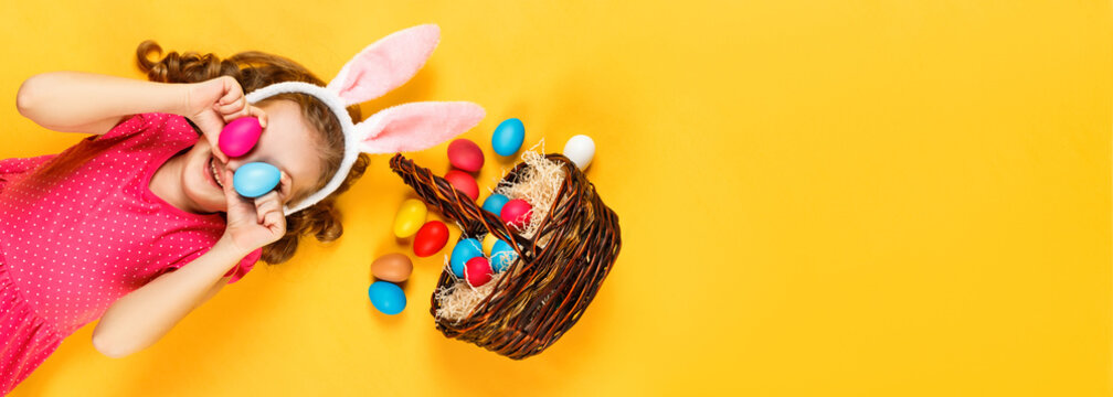 Top view on a happy merry child in bunny ears and a basket with Easter colored eggs. Little girl lies on the floor on a yellow background. Copy space