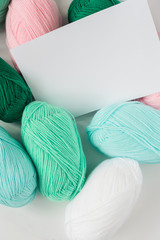 white paper sheet on acrylic soft pastel colored wool yarn thread skeins heap on white background, top view flat lay vertical stock photo image mockup with copy space for text