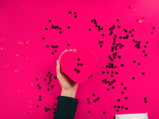 Obraz na płótnie Canvas Happy valentines day concept flat lay with red heart confetti on pink background and heart shaped gift box in female hands. Copy space