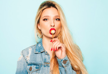 Portrait of young beautiful hipster bad girl in trendy jeans summer clothes and earring in her nose.Sexy carefree smiling blond woman posing in studio.Positive model licking round sugar candy