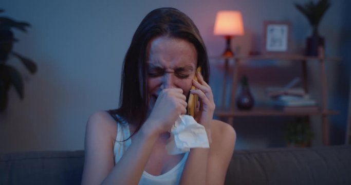 Close up of sad girl crying and shauting while talking on smartphone. Unhappy woman cant beliave bad news hearing during phone call while sitting on sofa . Concept of emotional conversation.