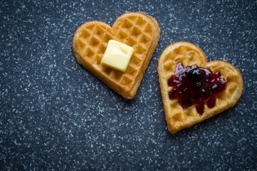 Pair of love heart-shaped waffles topped with berry jam and butter on a grey stone background