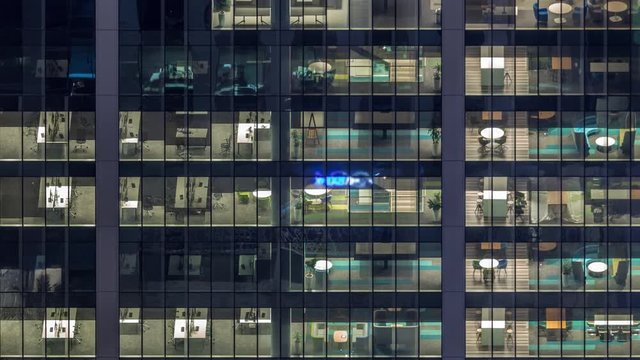 Office building exterior during late evening with interior lights on and people working inside night timelapse. Aerial close up view from above with many illuminated windows. Pan right