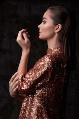 Young pretty woman in a sparkling dress with sequins stands with face in profile. Lady posing with her hands and elegant fingers on a black background. Fashion portrait of a girl