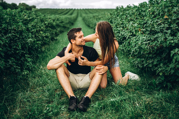 A smiling couple, woman with long hair and man sitting between currant bushes and hugging. Background of green currant plantations