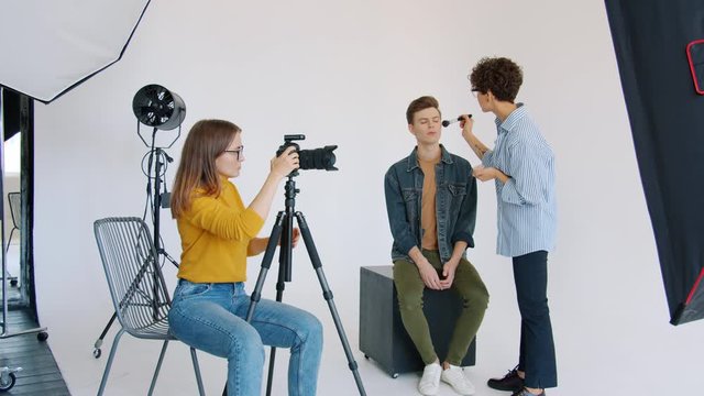 Photographer is adjusting digital camera while male model is working with make-up artist in studio getting ready for photoshoot. People and occupation concept.