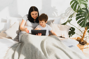 Beautiful smiling mother and son playing with digital tablet laying on bed.