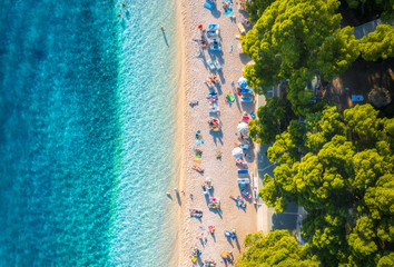 Aerial view of sandy beach with colorful umbrellas, swimming people, clear sea bay with transparent blue water and green trees at sunset in summer. Travel in Croatia, Adriatic sea. Top view. Seascape