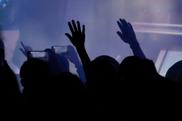 Hand with a smartphone records live music festival, Taking photo of concert stage. Party at nightclub with crowd of people raising hands with phones.