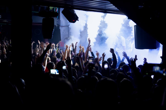 Audience With Hands Raised At A Music Festival With Colored Lights Streaming Down From Above The Stage. Rock Or Rap Concert With Many People.