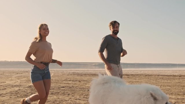 A happy smiling young couple man and woman are running with a dog samoyed on the lake in summer