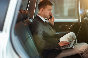 young businessman works in his laptop having business phone conversation while sitting in his car...