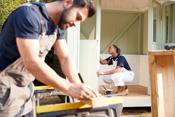 Male Carpenter With Female Apprentice Measuring Wood To Build Outdoor Summerhouse In Garden