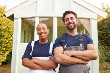 Male Carpenter With Female Apprentice Standing In Front Of Finished Outdoor Summerhouse In Garden