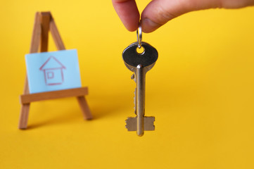 Drawing of a house on a wooden easel, nearby are the keys on a yellow background. The concept of buying or renting a house, apartment.