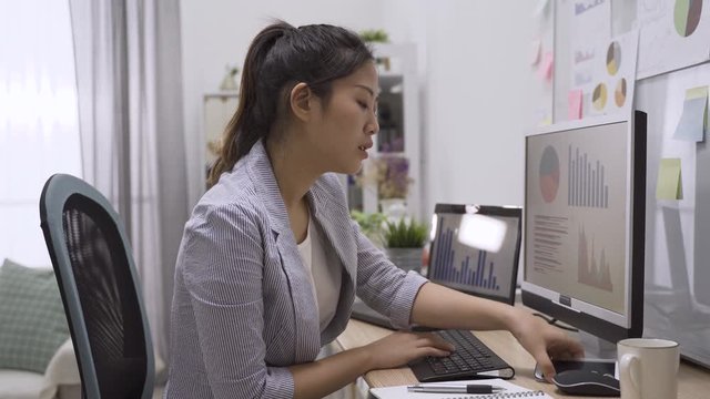 asian japanese woman working on computer at modern office while talking on mobile phone and discussing on chart. young girl worker having conversation on cellphone and pointing pc monitor indoors.