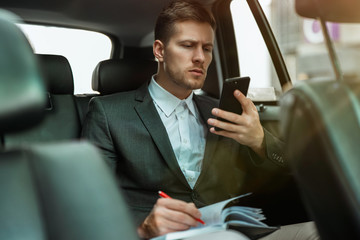 successful businessman takes notes to planner from his smartphone while riding on back seat of car on way to meetting with partners, multitasking concept