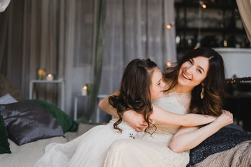 Mother and daughter hugging and cuddling in bed. Pretty little girl and beautiful woman lie together. Girls in lace dresses playing in decorated room. Family weekend, beauty day, having fun, love.