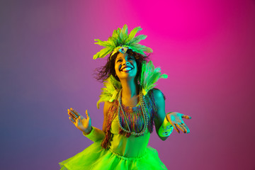 Obraz premium Crazy. Beautiful young woman in carnival, stylish masquerade costume with feathers dancing on gradient background in neon light. Concept of holidays celebration, festive time, dance, party, having fun