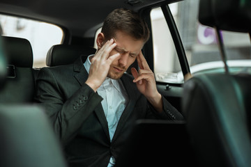 young businessman suffering extreme headache while riding on back seat of car showing like sign, looks miserable, healthcare and medical concept