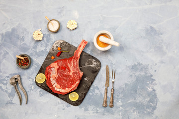 Raw Tomahawk steak on slate tray on light blue concrete background with spices and ingredients for...