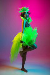 Happiness. Beautiful young woman in carnival, stylish masquerade costume with feathers dancing on gradient background in neon light. Concept of holidays celebration, festive, dance, party, having fun.