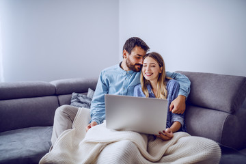 Cheerful cute caucasian couple sitting on sofa covered with blanket, snuggling and using laptop for...