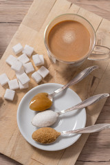 Fototapeta na wymiar Cukier w kostkach, cukier trzcinowy, cukier biały sypki oraz miód gryczany na łyżeczce leży obok szklanki z kawą z mlekiem.