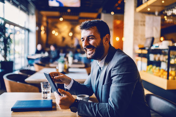 Young smiling caucasian bearded successful businessman sitting in cafe and using tablet for reading...