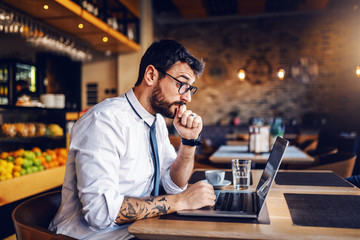 Young focused Caucasian bearded businessman with tattoo and eyeglasses in shirt and tie sitting in cafe and reading important e-mail from client.