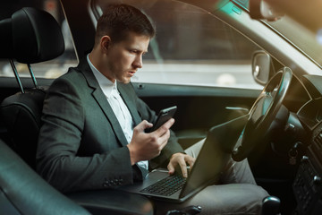 young businessman works in his car using his laptop and smartphone getting ready for important...