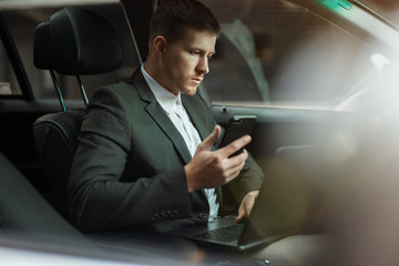 young businessman works in his car using his laptop and smartphone getting ready for important...