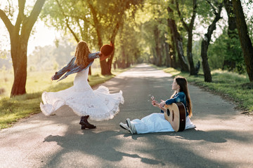 A girl in country-style clothes in a long dress dances on the road with a guitar at sunset. Happy childhood.