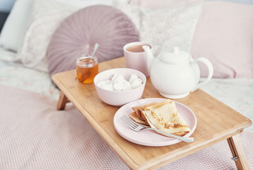Breakfast in bed in hotel room. Accommodation. Breakfast in bed with tea cup with pancakes on tray on bed background top view. Copy Space. Romantic valentine's day breakfast. Cozy breakfast Maslenitsa