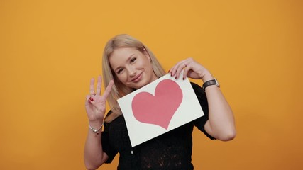 Beautiful blonde girl in black dress on yellow background stylish cool woman shows three fingers holding piece of paper with red heart